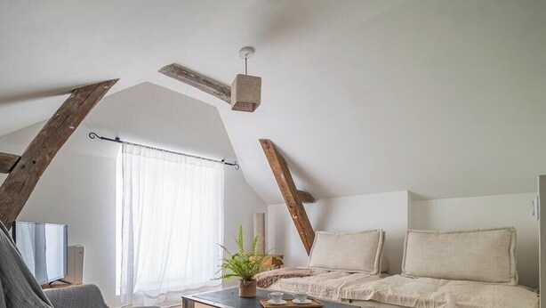 Dormitorio tipo loft con vigas de madera a la vista, una cama, un escritorio y un ventanal con cortinas.