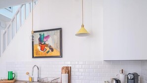 Una cocina con armarios blancos, un cuadro colgado, una lámpara colgante amarilla y una escalera.