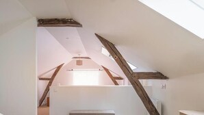 Una habitación con techo inclinado, vigas de madera, tragaluces y una lámpara colgante.