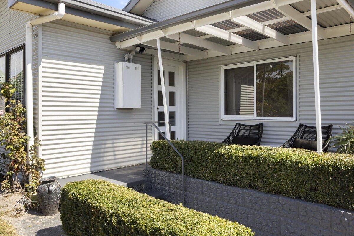 A residential exterior with a covered porch, a window with blinds, a white door, and a small garden area with a potted plant and a decorative urn.