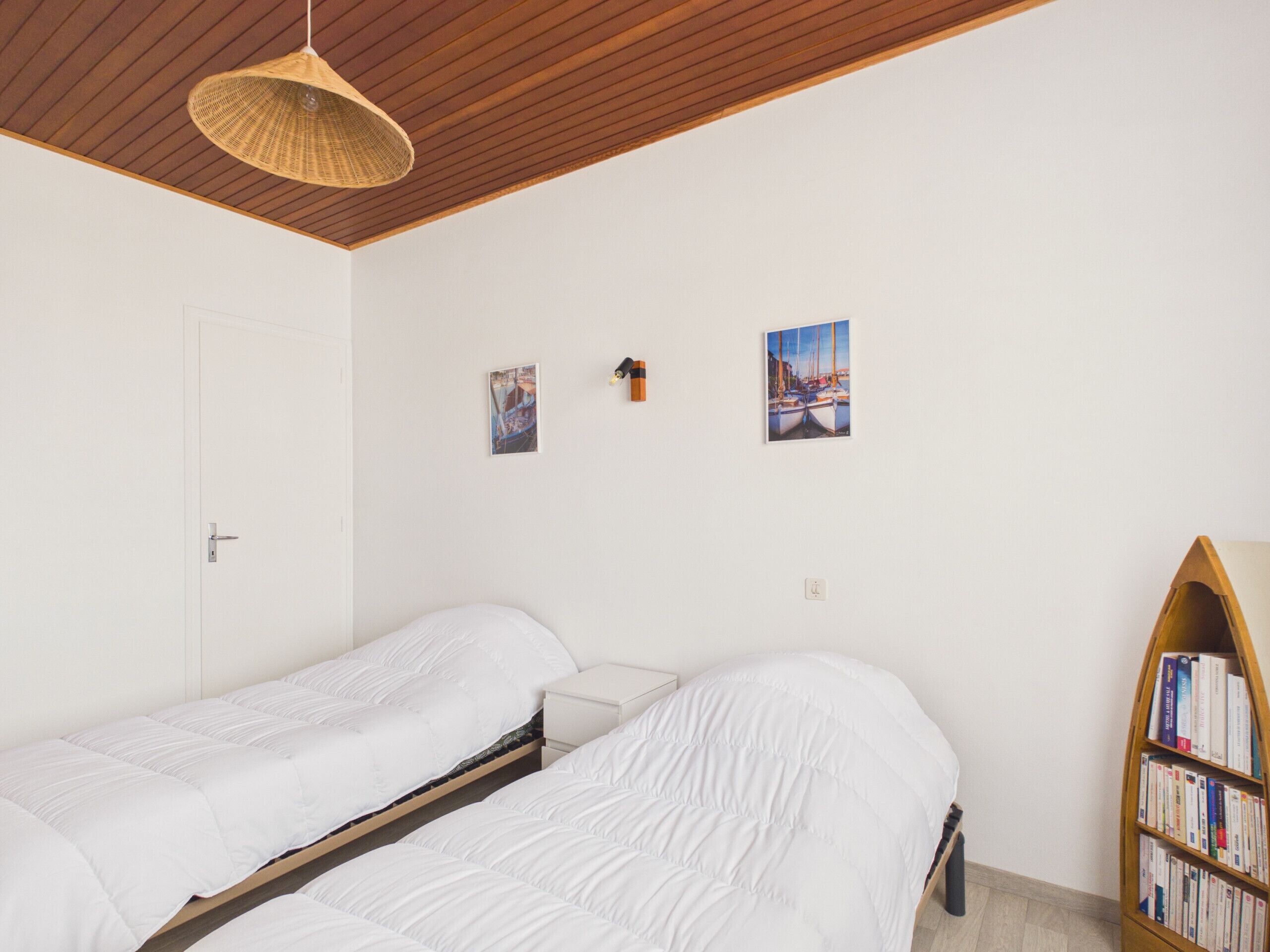 Una habitación con dos camas individuales, techo de madera, una estantería y dos cuadros colgados.