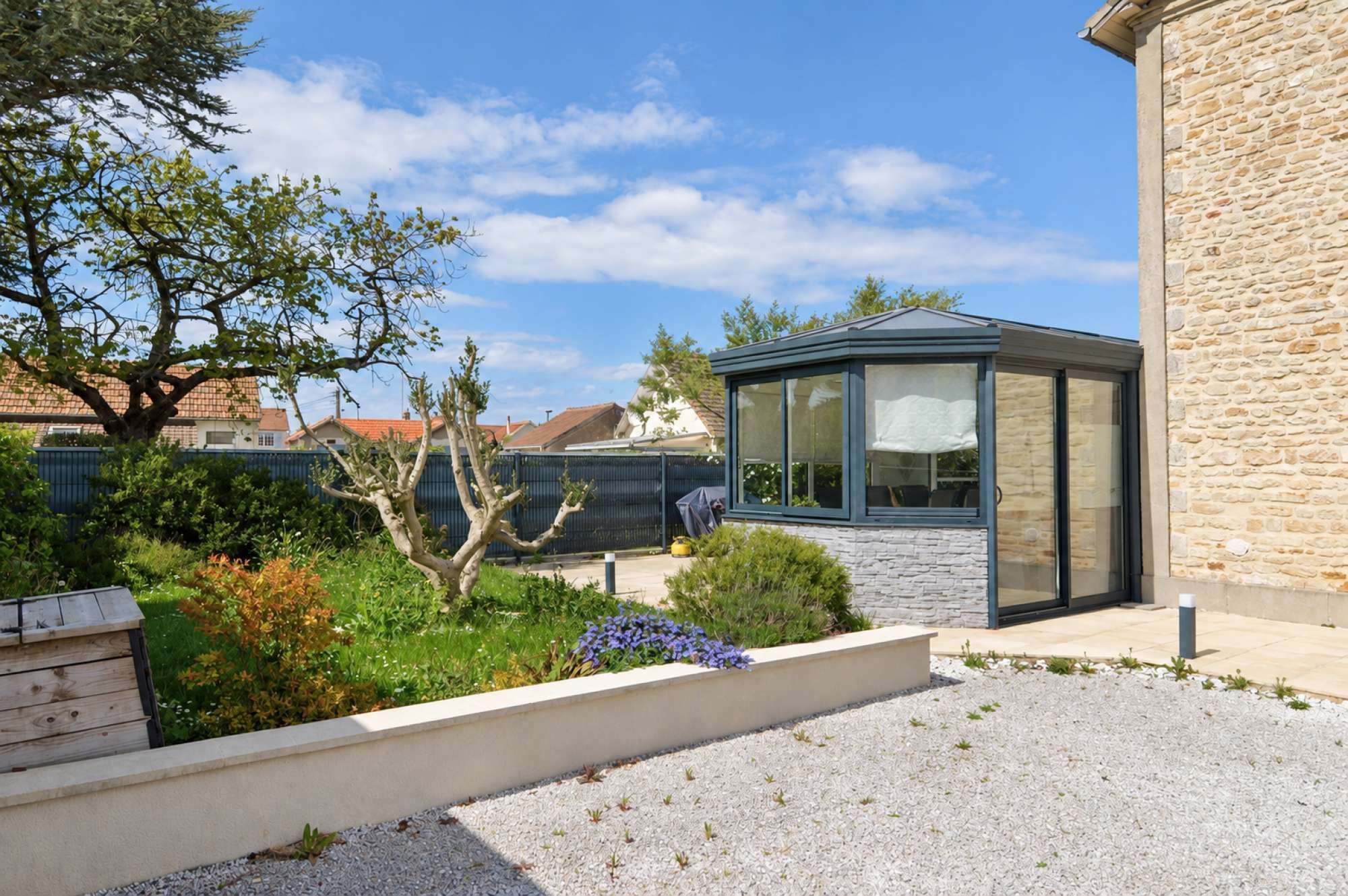 A modern house with a conservatory, a garden with various plants, and a stone wall.