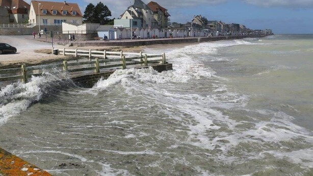 Een kustpromenade met gebouwen, een houten steiger en golven die op het strand breken.