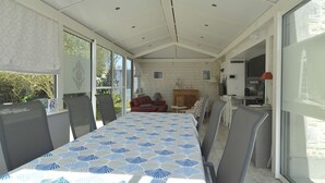 Een ruime kamer met een lange tafel, meerdere stoelen, een open haard en een keukenhoek.