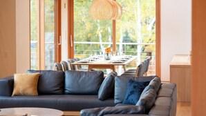 A modern living room with a dark leather sofa, a dining area with a wooden table, and a large window with a view of trees. - Ro-An (Kutchan)