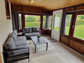 Une pièce avec un plafond en bois, un canapé gris, une petite table avec une plante et de grandes fenêtres avec des rideaux.