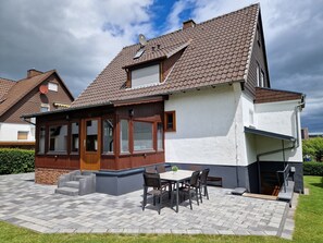 A house with a tiled roof, a covered patio with a table and chairs, and a small garden area.