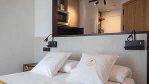 Una cama bien hecha con ropa blanca y almohadas, una mesita de noche, una lámpara de pared y un espejo sobre la cama.
