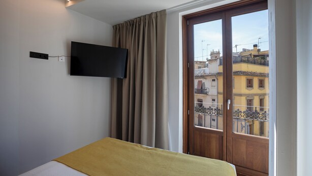 A bedroom with a bed, a television mounted on the wall, and a window with a view of buildings. - Mon Suites Platería (Valencia)