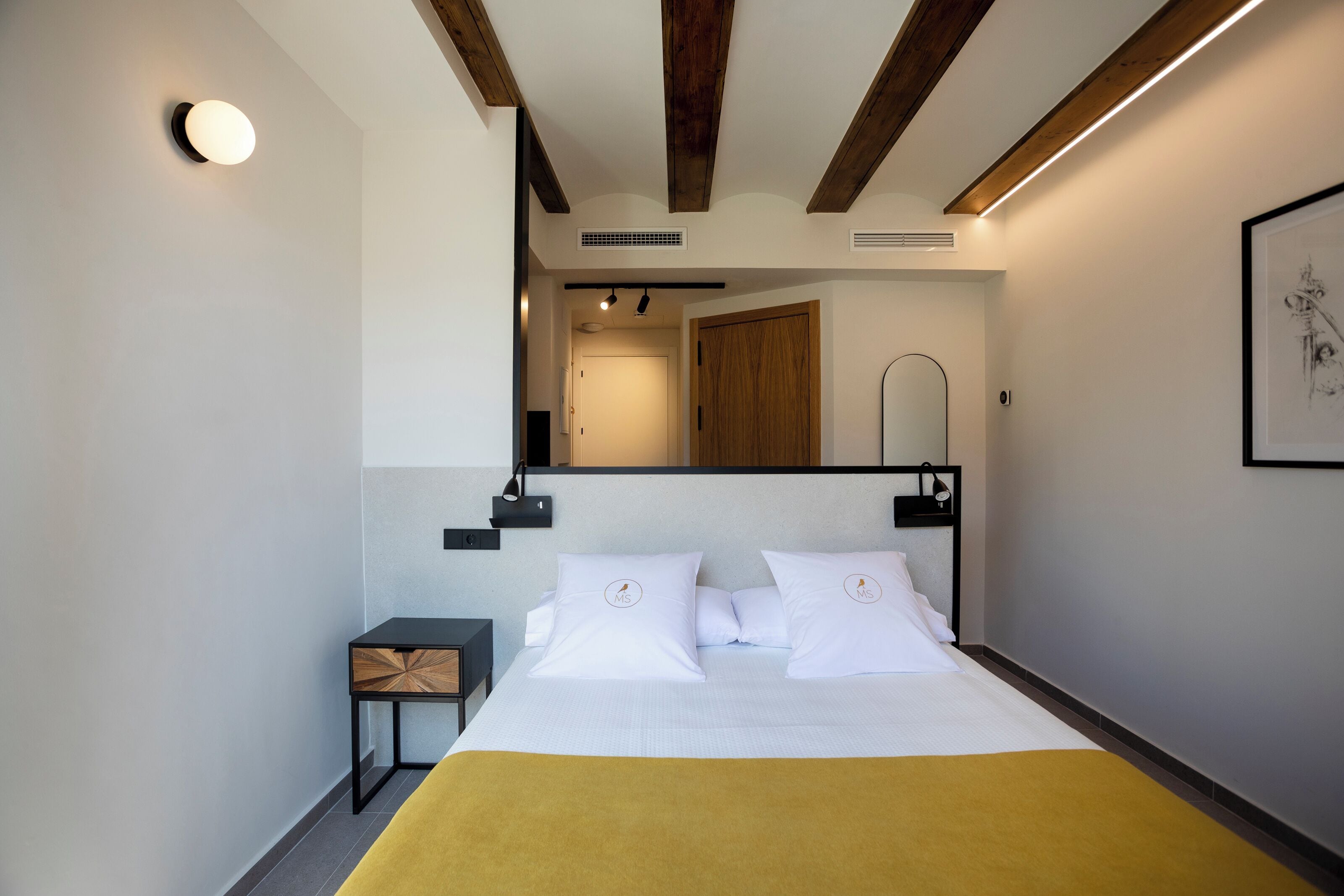 A modern hotel room with a large bed, a bedside table, a mirror, and a framed artwork on the wall.