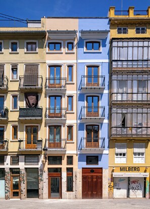 A row of multi-story buildings with balconies, a shop on the ground floor, and a visible sign reading 'MILBERG'. - Mon Suites Platería (Valencia)