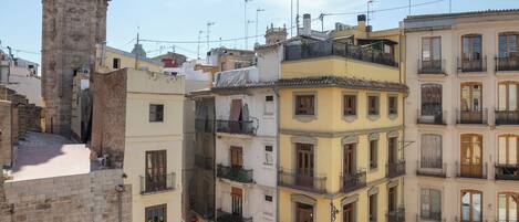 Una torre histórica con una campana, edificios de varios pisos adyacentes con balcones, y una zona peatonal debajo.