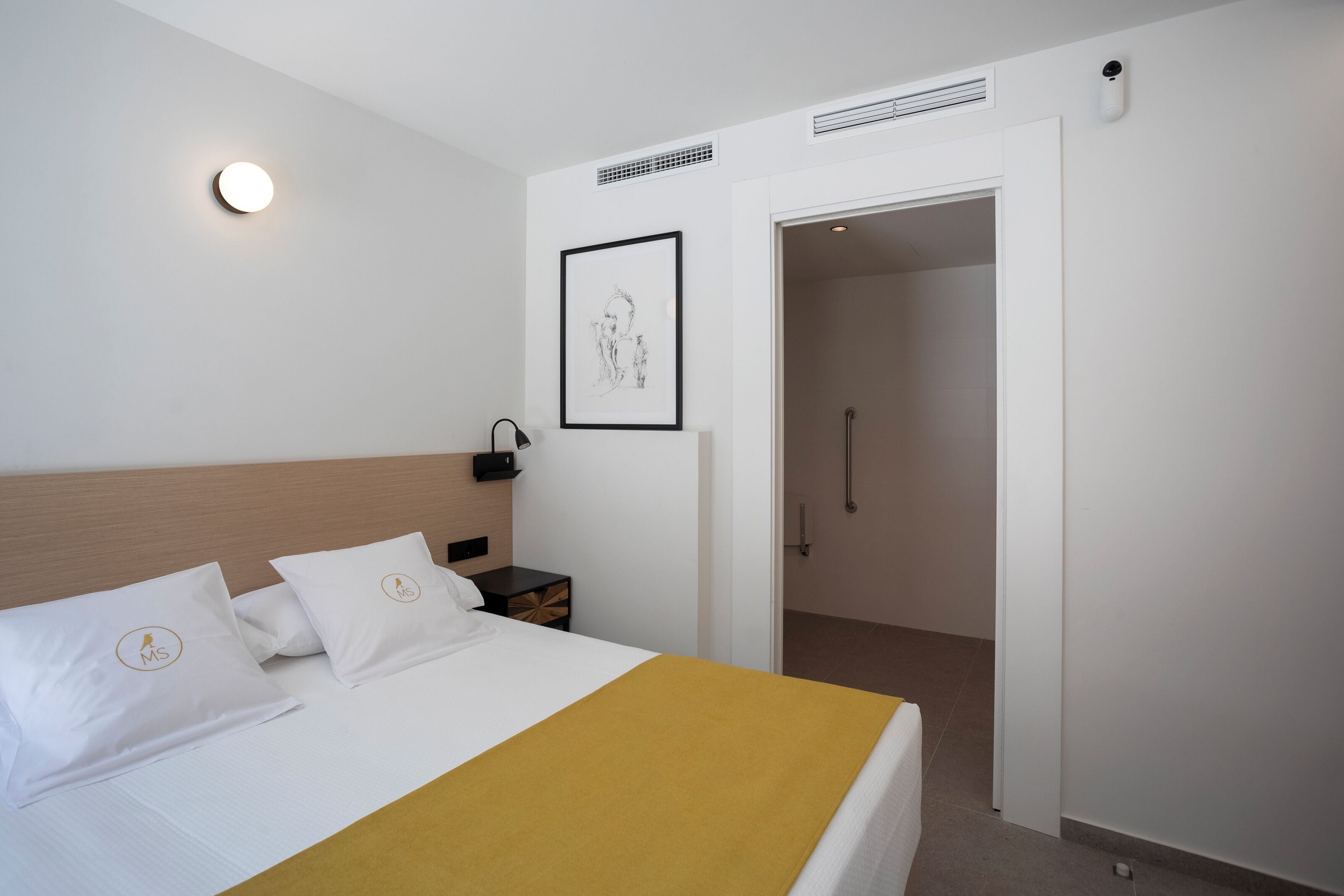Habitación de hotel con cama, mesita de noche, lámpara de pared, cuadro enmarcado y puerta abierta hacia un baño.