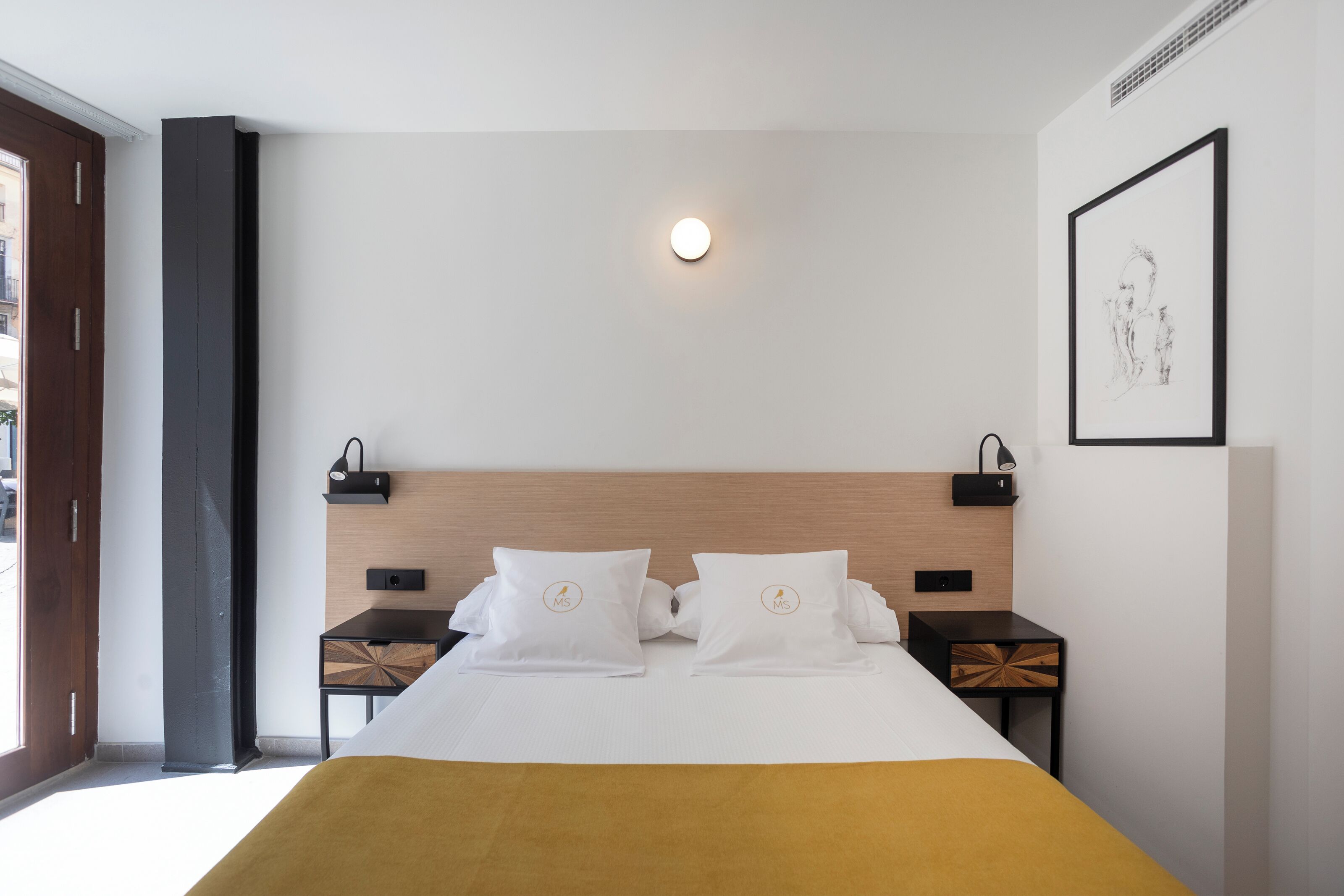 Quarto de hotel moderno com cama, mesas de cabeceira, luminária fixada na parede e uma obra de arte emoldurada.