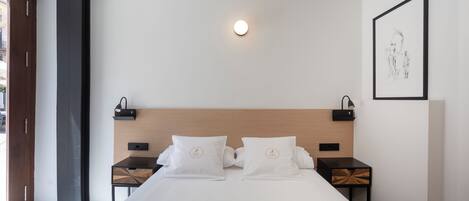 Una habitación de hotel moderna con cama, mesitas de noche, una lámpara de pared y una obra de arte enmarcada.