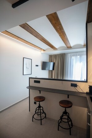 A modern room with a wooden ceiling, a flat-screen TV mounted on the wall, a window with curtains, and two stools at a counter. - Mon Suites Platería (Valencia)