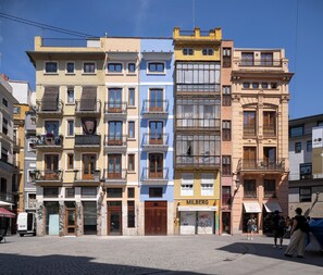A row of multi-story buildings with balconies, a visible storefront, and a cobblestone street. - Mon Suites Platería (Valencia)