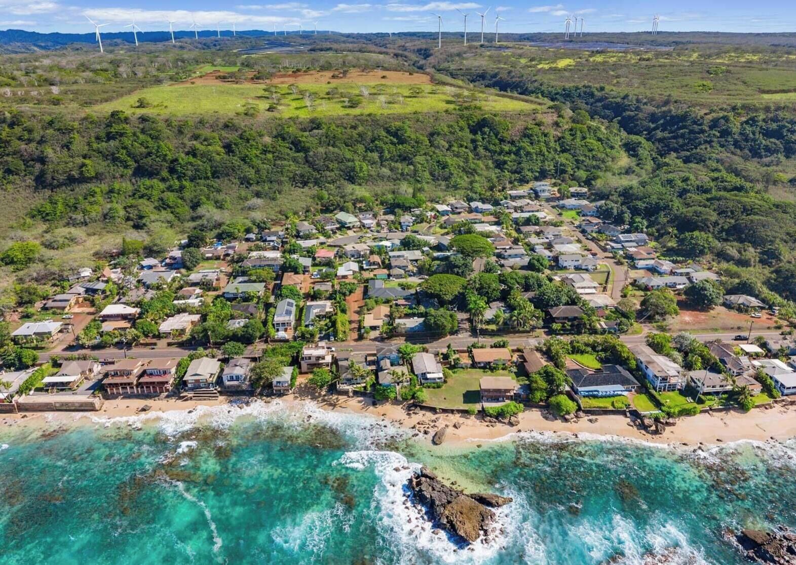 An aerial view of a coastal residential area with houses, a beach, and wind turbines in the background.