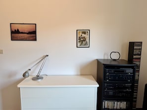 Ein Zimmer mit weißem Schreibtisch, einer Lampe, einem Bücherregal mit CDs und zwei gerahmten Bildern an der Wand.