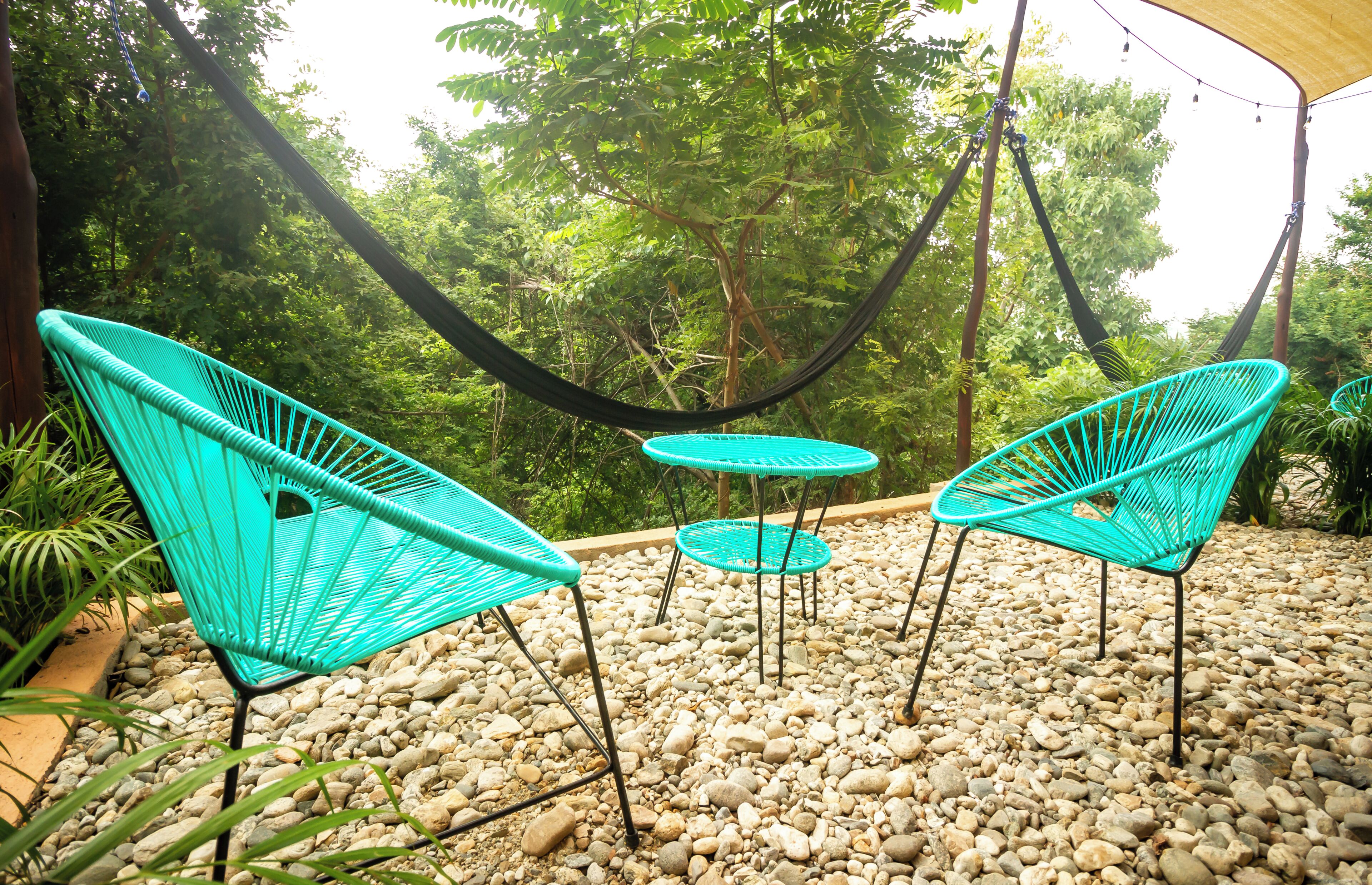 A hammock suspended between trees, surrounded by turquoise chairs and a small round table on a gravel surface.