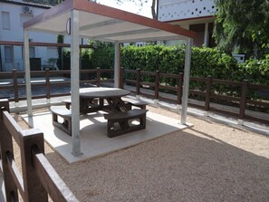 An outdoor covered picnic area with tables and benches, surrounded by a fence and greenery. - Comfortable stay with balcony and garden (Lignano Sabbiadoro)