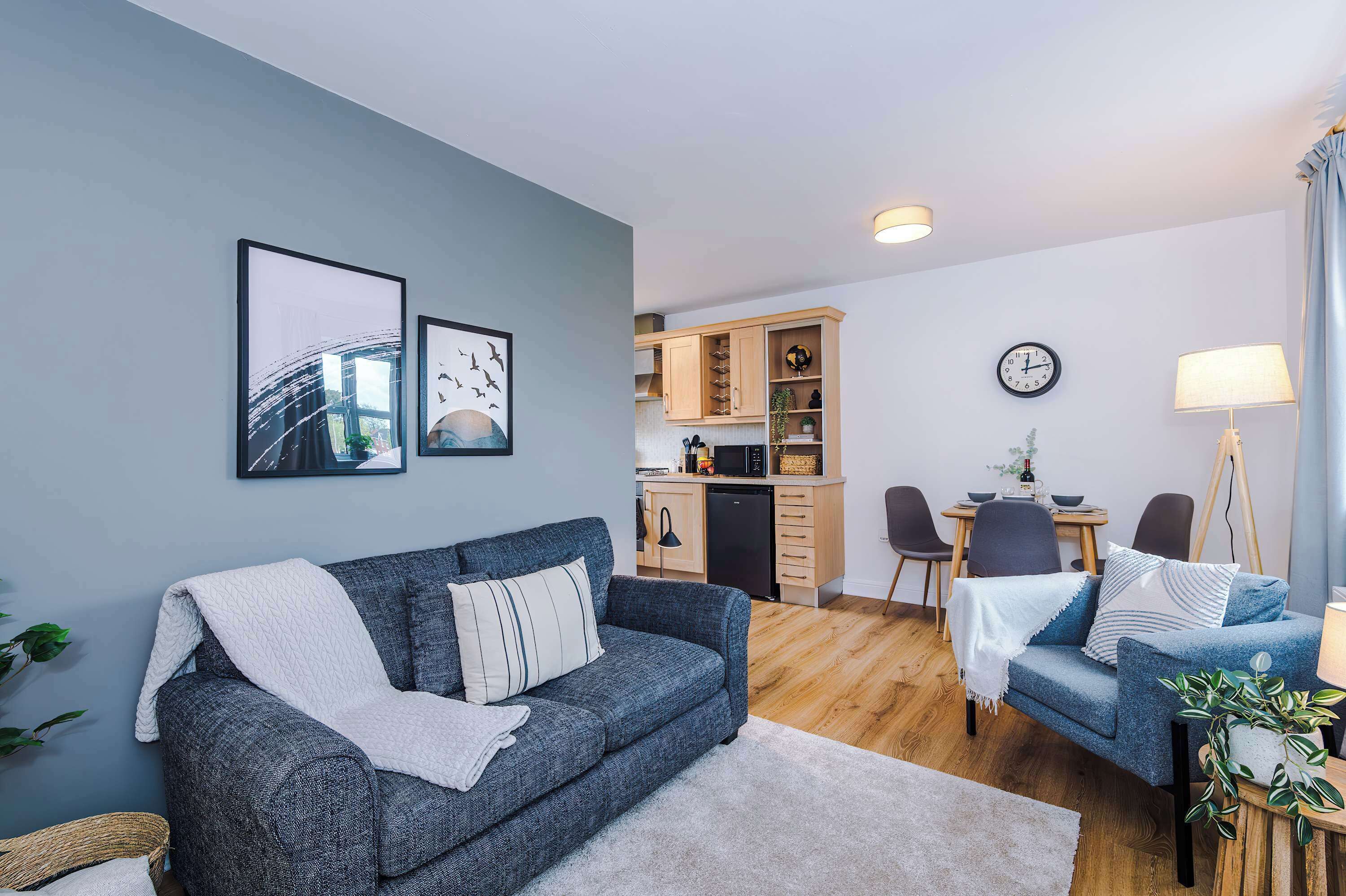 A modern living room with a grey sofa, a dining area with a table and chairs, and a kitchen area with wooden cabinets and a microwave.