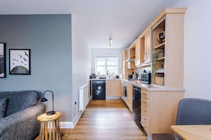A compact kitchen with wooden cabinets, a washing machine, and a dining area with a table and chairs. - Stunning 1 Bed Flat, Perfect for Business in Crewe (Cheshire East)