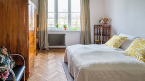 Ein Schlafzimmer mit einem Bett, einem Holzschrank, einem Stuhl, einem Fenster mit Vorhängen und einem Heizkörper.