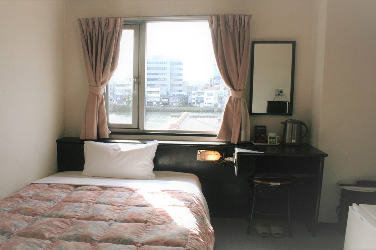 A bedroom with a bed, a desk, a chair, a window with curtains, and a city view.