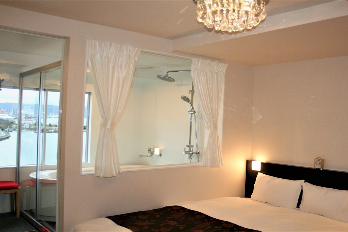 Habitación de hotel con cama, ducha, ventana con cortinas y vista a la ciudad.