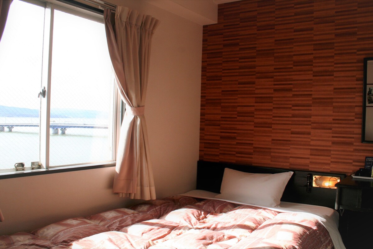 Une chambre d’hôtel avec un lit, une fenêtre avec des rideaux, un mur en bois, une table de chevet avec une lampe et une vue sur un pont.
