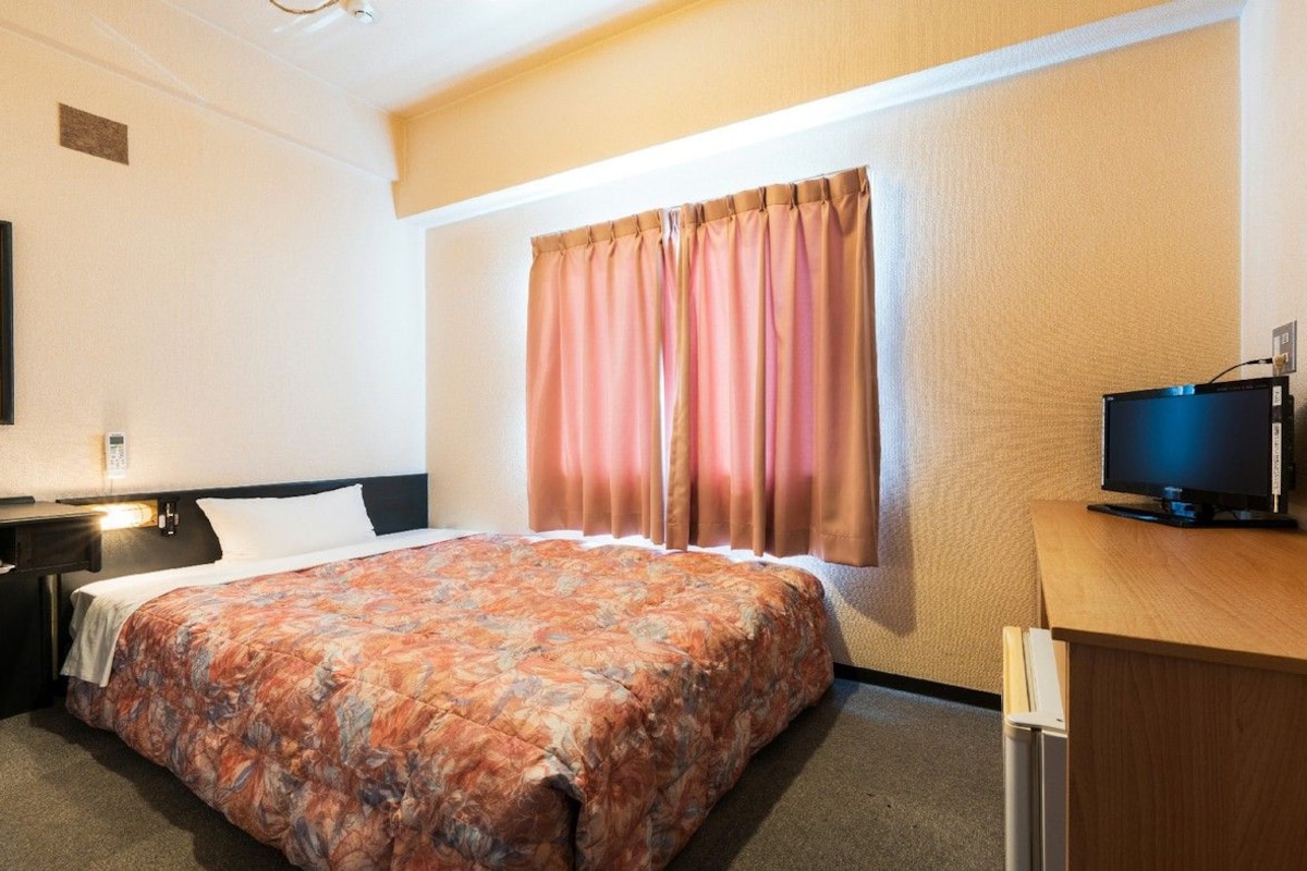 Una habitación de hotel con cama, escritorio, televisor y ventana con cortinas.