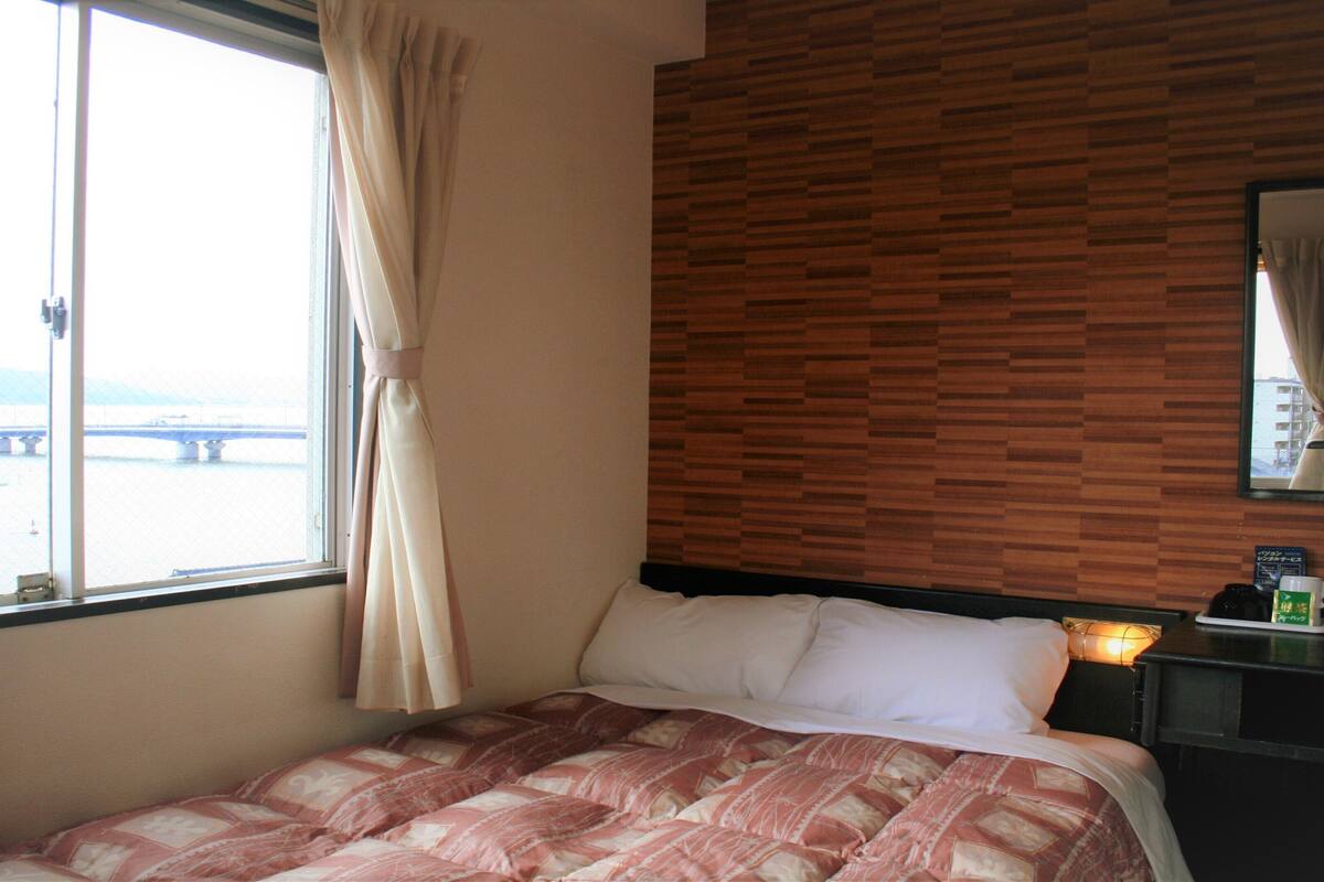 A hotel room with a bed, a nightstand, a window with curtains, and a view of a bridge and water.