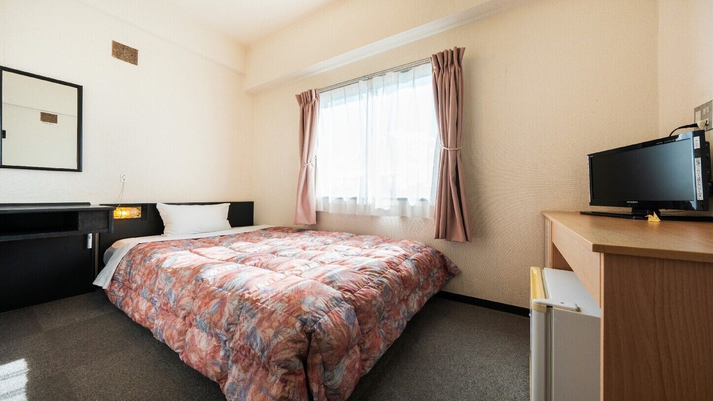 Une chambre d’hôtel avec un lit, un bureau avec une télévision, un miroir et une fenêtre avec des rideaux.