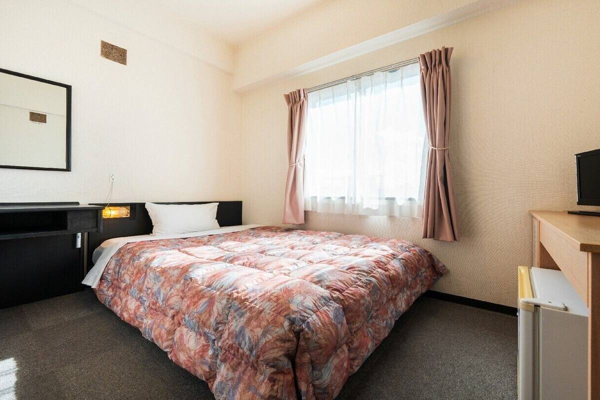 A hotel room with a bed, a desk with a television, a mirror, and a window with curtains.