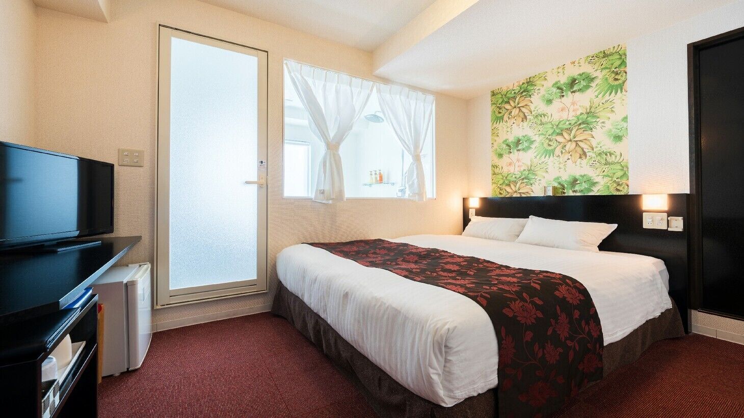 Une chambre d’hôtel avec un lit, une télévision, une fenêtre avec des rideaux et un mur orné d’un motif floral.
