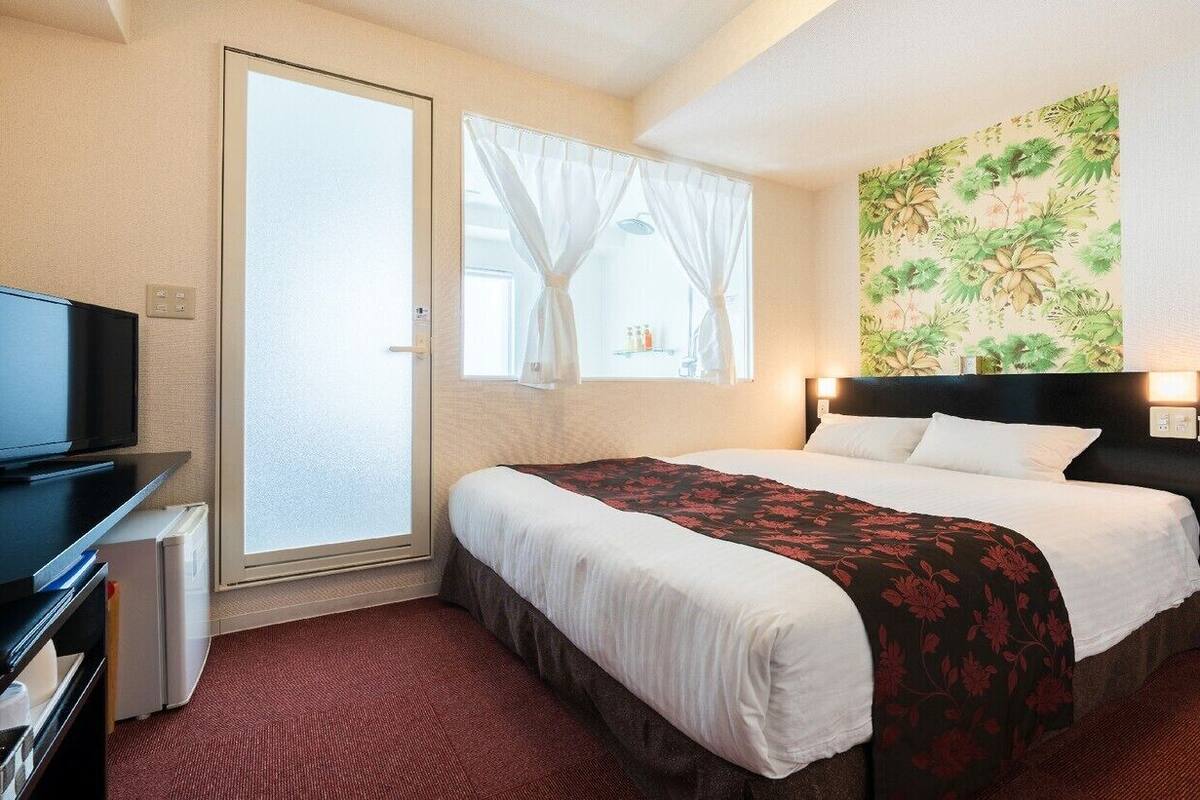 A hotel room with a bed, a television, a window with curtains, and a wall with a floral pattern.