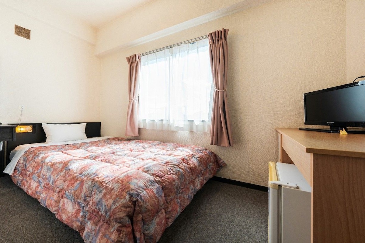 Una habitación de hotel con una cama, un escritorio con televisor, un espejo y una ventana con cortinas.