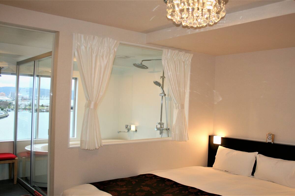 A hotel room with a bed, a shower, a window with curtains, and a view of a cityscape.