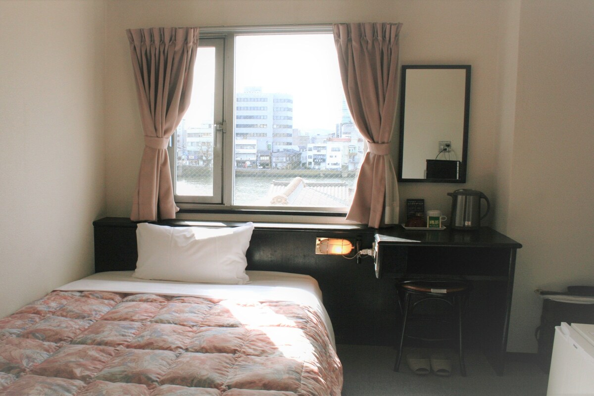 Un dormitorio con cama, escritorio, silla, ventana con cortinas y vistas a la ciudad.