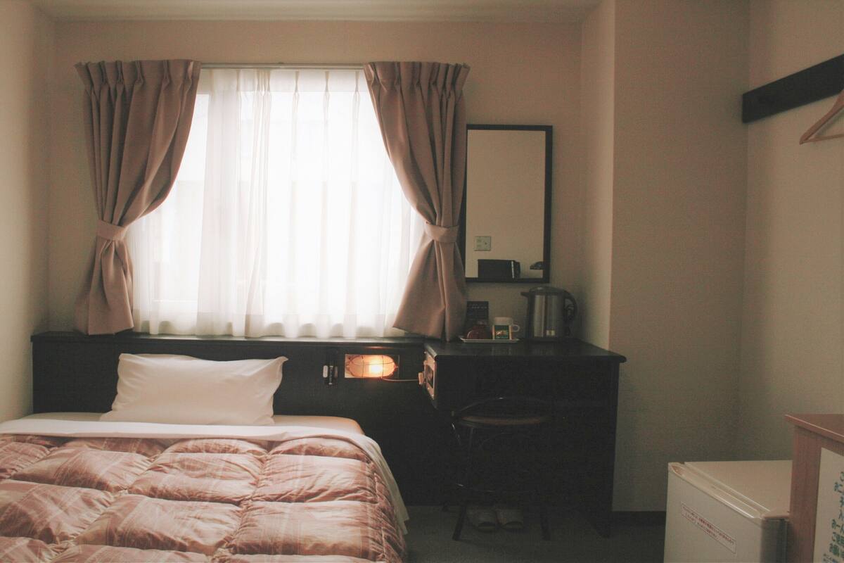 A hotel room with a bed, a desk with a lamp, a chair, and a small refrigerator.