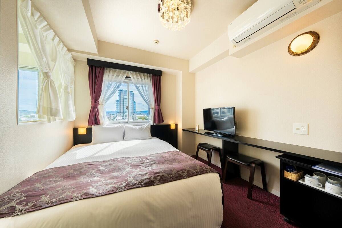 A hotel room with a bed, a desk with a TV, a chair, a small table, and an air conditioning unit.