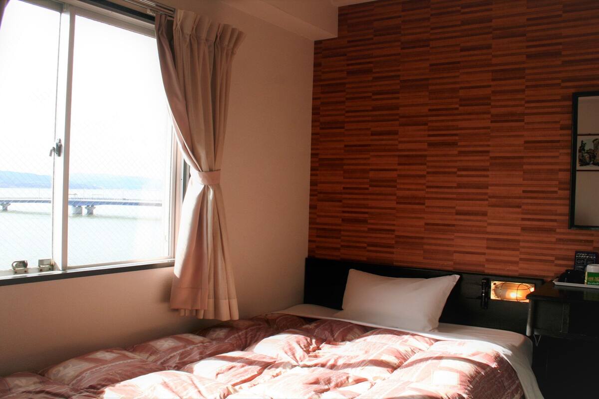 A hotel room with a bed, a window with curtains, a wooden wall, a bedside table with a lamp, and a view of a bridge.