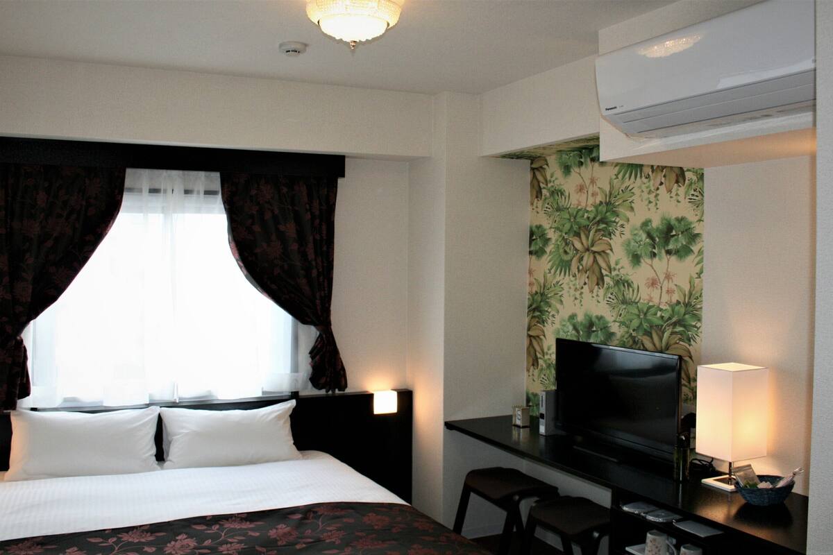 A hotel room with a bed, a desk with a TV, a chair, and an air conditioning unit.