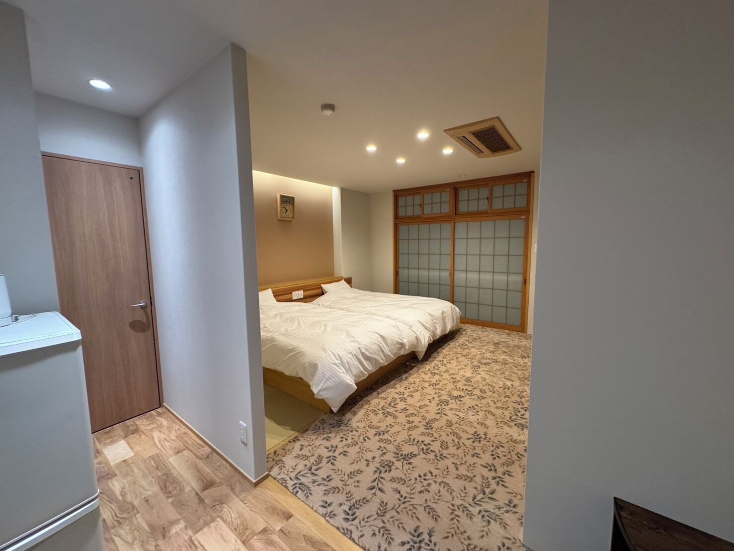 Un dormitorio con cama, un armario de madera, una mesita y una alfombra estampada.