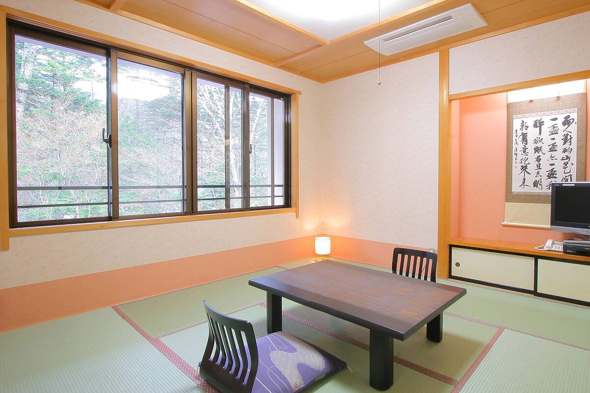 A traditional Japanese room with tatami flooring, a low table, chairs, a television, and a calligraphy piece on the wall.