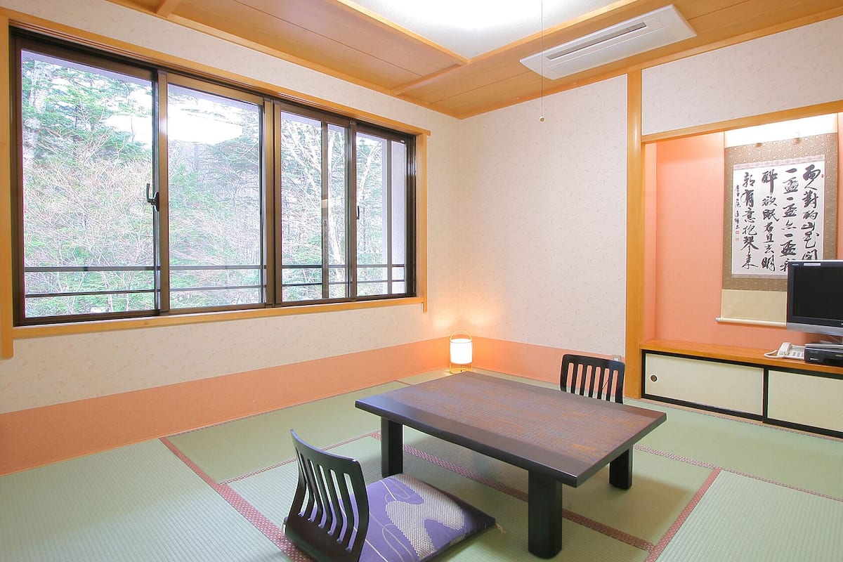 Una habitación japonesa tradicional con suelo de tatami, una mesa baja, sillas, un televisor y una obra de caligrafía en la pared.