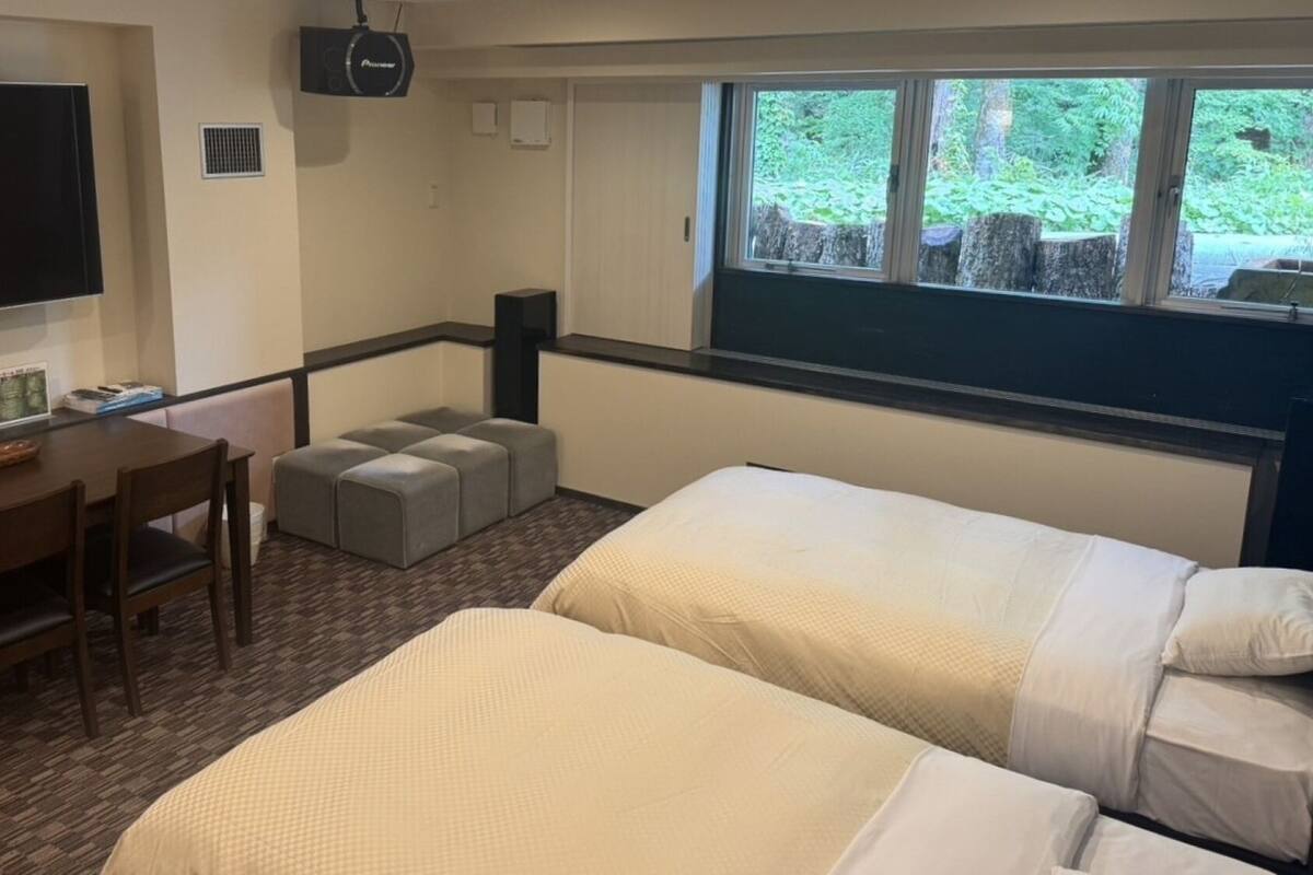A hotel room with two beds, a desk with chairs, a television, and a window with a view of greenery.