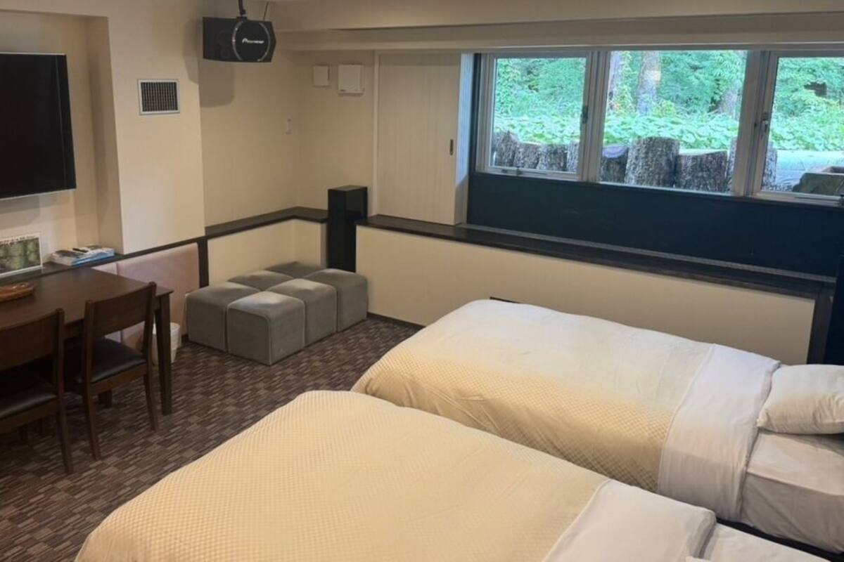 A room with two beds, a desk with chairs, a television, and a window with a view of trees.