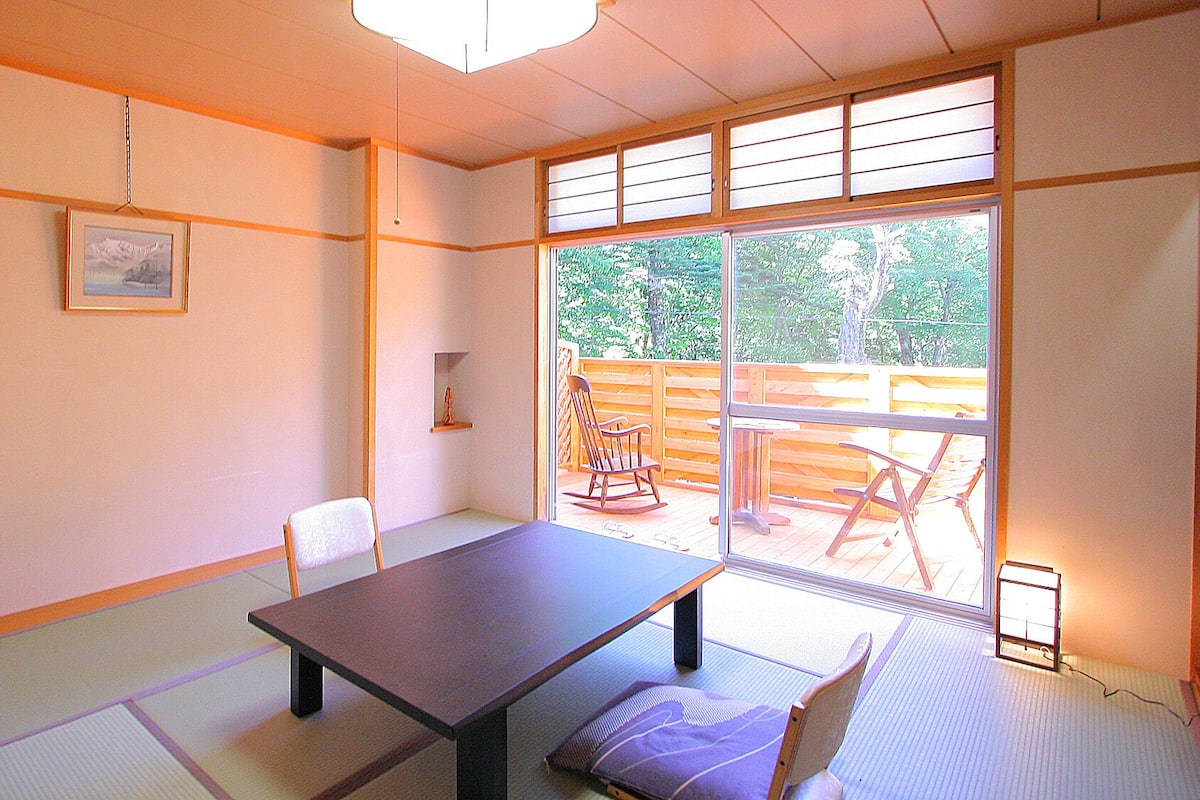 A room with tatami flooring, a low table, a chair, and a view of an outdoor deck.